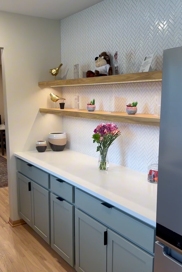 Covington kitchen counter with sage cabinets, herringbone backsplash, floating shelves, and fresh flowers