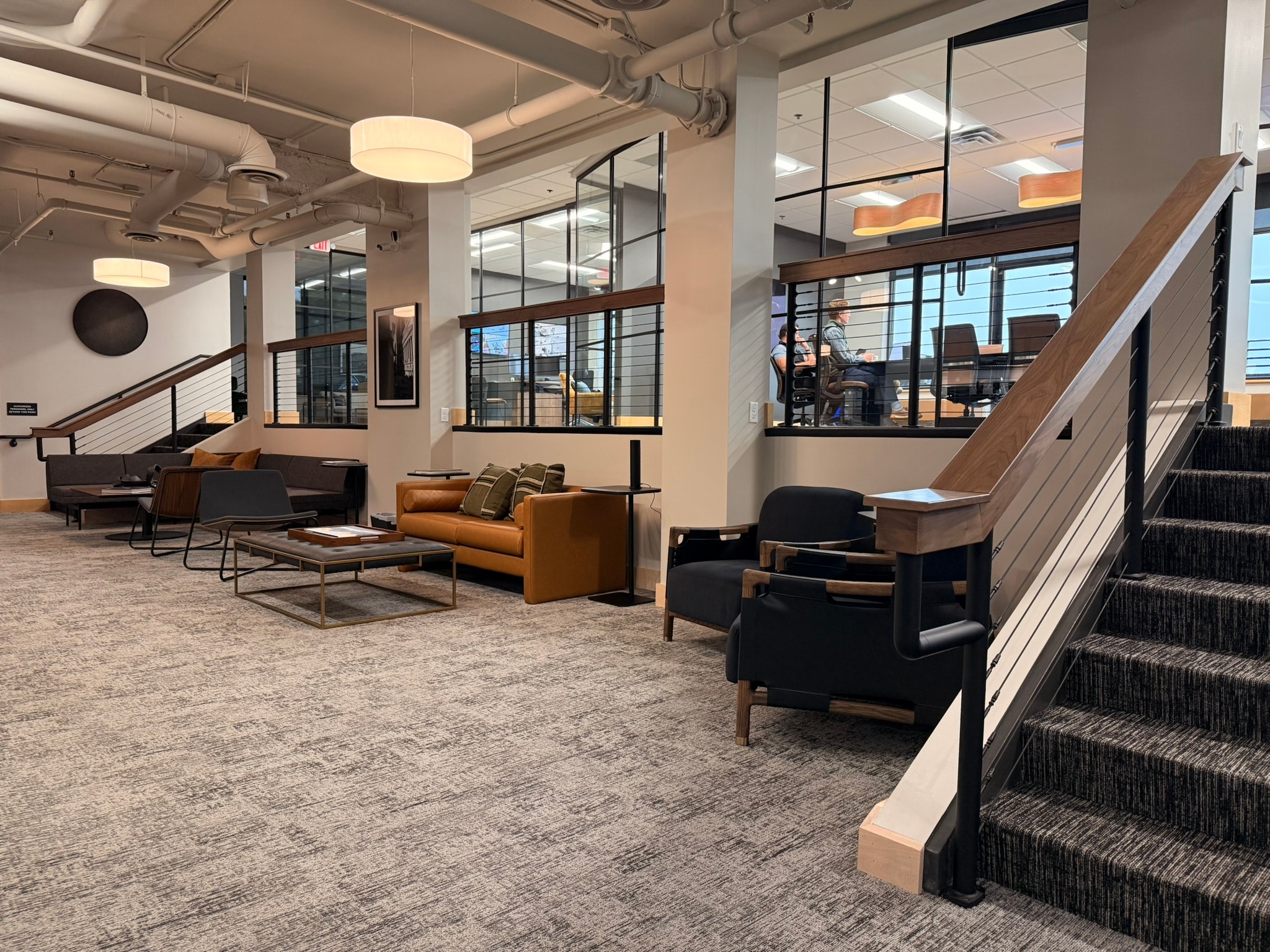SnowPine Private Wealth lower lounge with cognac leather sofa, cable-rail staircase, and glass office windows above