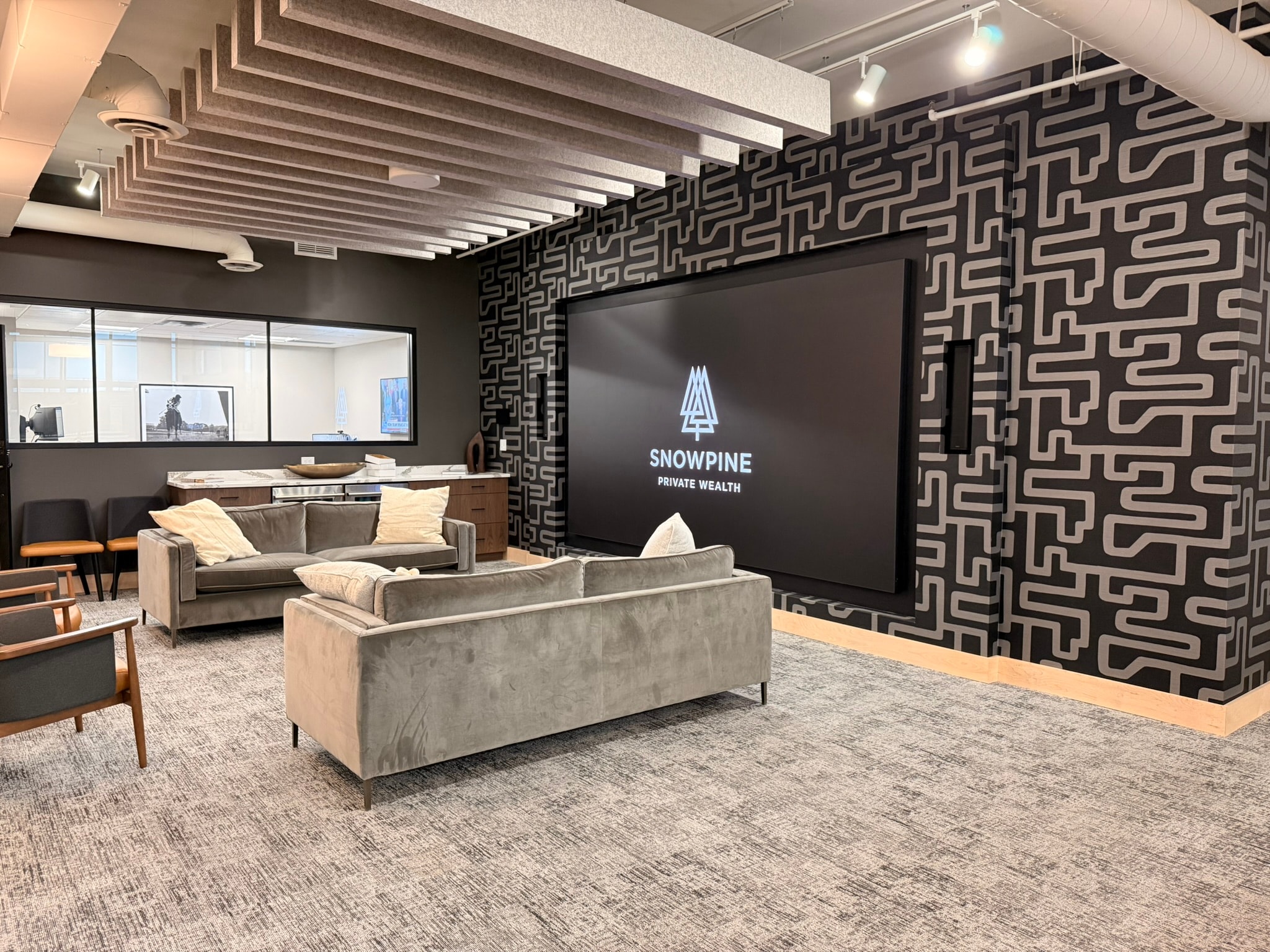 SnowPine Private Wealth main lounge with gray velvet sofas, geometric feature wall, and SnowPine logo on large display screen