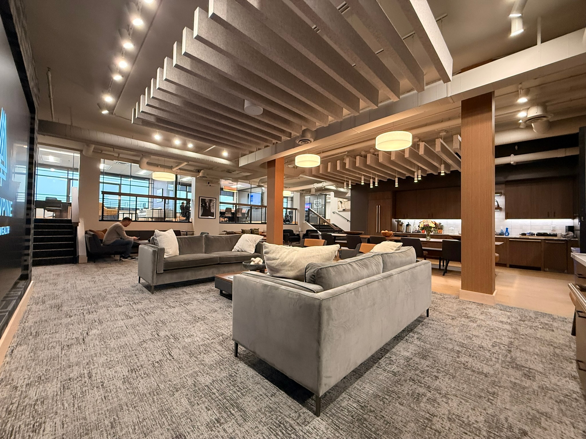 SnowPine Private Wealth main floor wide view with gray sofas, acoustic ceiling baffles, and kitchen zone beyond